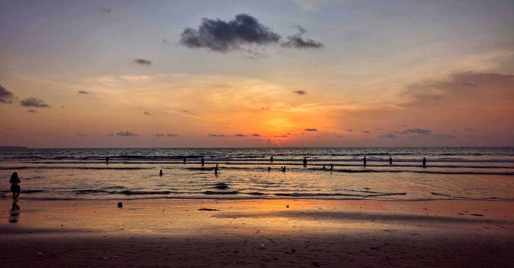 A wide Bali beach at sunset, the wet sand mirroring an orange sky and distant figures along the waterline.