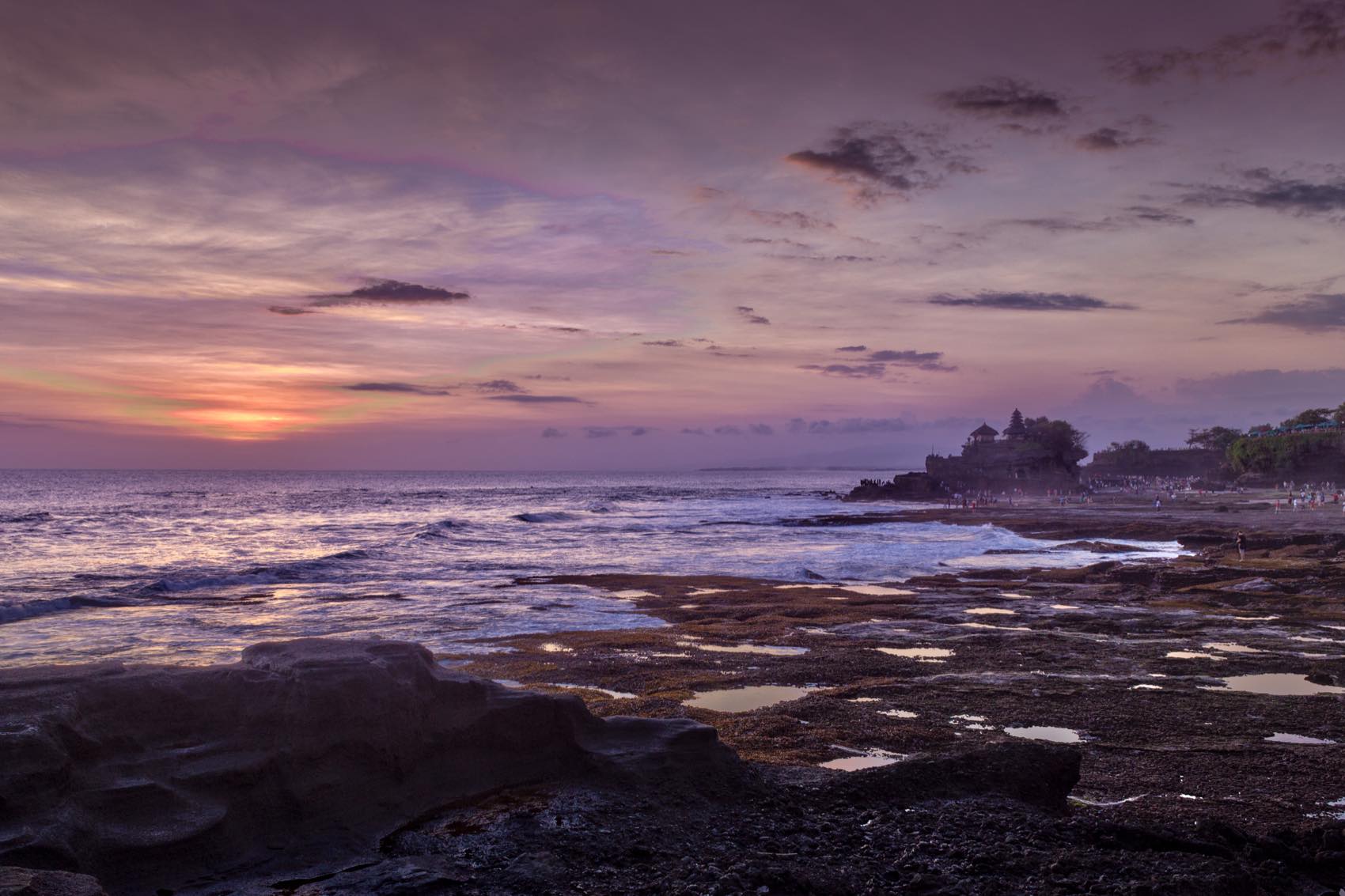 Tanah Lot sea temple at dusk, silhouetted on its rock above the tide with the sun low over the water.