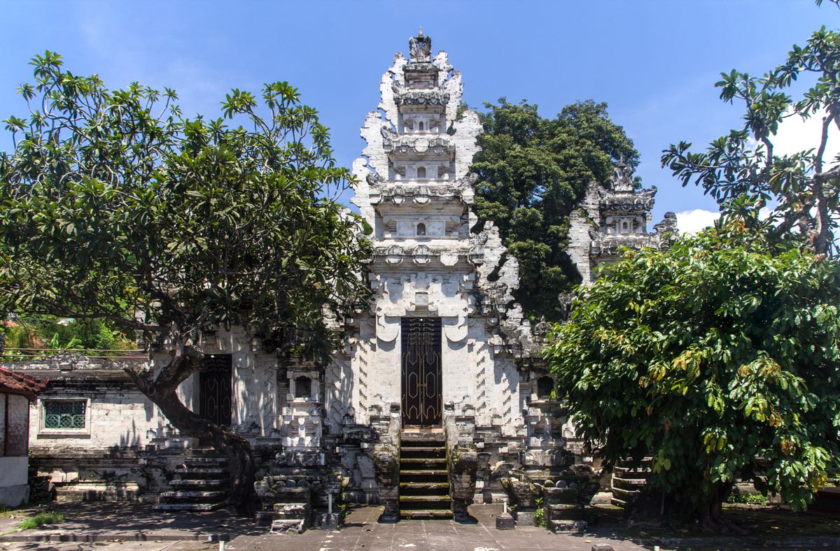 A Balinese temple gate in weathered pale stone, framed by frangipani and mango trees against a blue sky.